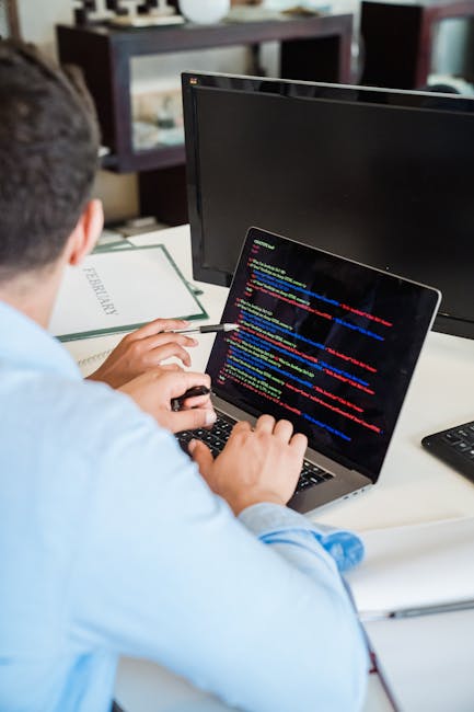 The image shows a person in a light blue shirt working at a white desk in an indoor office setting. They are using a laptop, which displays colorful lines of computer code on a dark background. The in